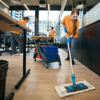 Team of cleaners in overalls and rubber gloves work in a coworking area, women use a mop, buckets, rags